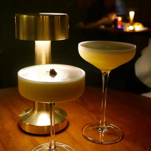 Two elegant cocktails in stemmed glassware with frothy tops, one under a gold table lamp.